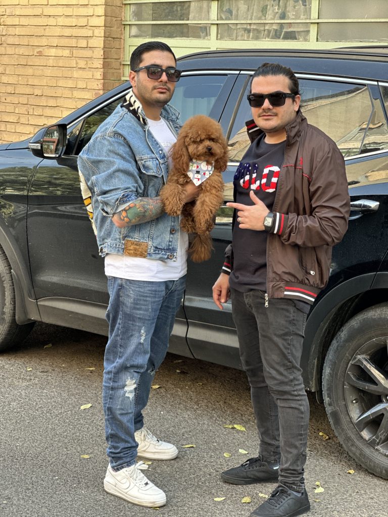 "Two people holding a medium-sized brown poodle in front of a car."